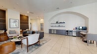 Located at 1614 Hill St in Grand Prairie, Texas, this contemporary lobby space offers a welcoming and well-appointed environment designed for both comfort and functionality. The single-floor layout provides a generous, open feel, with large-format light-toned tile flooring extending throughout and contributing to a clean, airy aesthetic.

The space is anchored by multiple distinct seating areas, including a central lounge furnished with leather sofas, accent chairs, and a patterned area rug. A standout architectural feature is a dramatic arched alcove framing a built-in display wall with floating shelves and gray cabinetry, complete with under-counter beverage refrigerators—ideal for a hospitality or residential amenity setting.

Tall ceilings amplify the sense of openness, while recessed lighting and strategically placed wall sconces create a warm, inviting ambiance. A textured, patterned accent wall in dark tones adds depth and visual interest, complemented by built-in banquette seating with leather cushions and modern side tables. A wall-mounted flat-screen TV and a sleek fireplace panel are integrated into a light wood feature wall, creating a cozy media area.

Floor-to-ceiling windows and glass entry doors along one side flood the space with natural light and offer views to the exterior. Curated artwork and decorative accessories complete the refined, modern atmosphere of this well-designed lobby.