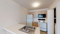 Located at 100 Windsor Cir in Jacksonville, North Carolina, this single-floor unit offers a straightforward, functional layout designed for comfortable everyday living. The kitchen is efficiently arranged with light wood cabinetry, white appliances including a full-size refrigerator, electric range, and built-in microwave, along with tile flooring and a double-basin sink set into a counter peninsula that separates the kitchen from the rest of the living space. A washer and dryer area is also accessible from the kitchen. The living area features neutral wall tones and plush wall-to-wall carpet that extends throughout the bedrooms, lending a warm and cohesive feel to the space. The bedrooms are bright and airy, benefiting from windows with blinds that allow natural light to filter in; one bedroom includes a closet and multiple windows overlooking the outdoors. Two bathrooms serve the unit — one features a pedestal sink and a tub/shower combo with clean white surrounds, while the second offers a soaking tub and separate towel and shower rod fixtures with tile flooring. The overall design is clean, neutral, and move-in ready, providing a practical and comfortable living environment.