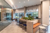 This single-floor amenity and lobby space showcases a thoughtfully curated modern design that seamlessly blends natural materials with contemporary architecture. Upon entry, large floor-to-ceiling glass doors and expansive windows flood the space with natural light, offering open views of the surrounding streetscape. The reception area features a sleek dark-topped counter framed by light wood cladding, backed by striking vertical slats of natural pine that add warmth and texture to the environment.

Geometric linear LED lighting runs across the ceiling in angular patterns, creating a dynamic visual element that ties the space together. Large-format tile flooring in a neutral palette extends throughout, grounding the space with a clean, polished aesthetic.

A fully glass-enclosed conference room serves as a focal point, featuring a gabion stone accent wall, patterned flooring, and modern pendant lighting above a dark conference table surrounded by burnt-orange chairs. The transparency of the glass walls maintains visual connectivity with the broader space.

Seating areas are strategically distributed throughout, including built-in benches with orange cushions and marble-topped side tables near the entrance, as well as a window-facing lounge with colorful upholstered benches and curated accent furniture. Lush indoor plants are incorporated throughout, reinforcing a welcoming, organic atmosphere that balances the industrial and natural design elements.