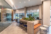 This single-floor amenity and lobby space showcases a thoughtfully curated modern design that seamlessly blends natural materials with contemporary architecture. Upon entry, large floor-to-ceiling glass doors and expansive windows flood the space with natural light, offering open views of the surrounding streetscape. The reception area features a sleek dark-topped counter framed by light wood cladding, backed by striking vertical slats of natural pine that add warmth and texture to the environment.

Geometric linear LED lighting runs across the ceiling in angular patterns, creating a dynamic visual element that ties the space together. Large-format tile flooring in a neutral palette extends throughout, grounding the space with a clean, polished aesthetic.

A fully glass-enclosed conference room serves as a focal point, featuring a gabion stone accent wall, patterned flooring, and modern pendant lighting above a dark conference table surrounded by burnt-orange chairs. The transparency of the glass walls maintains visual connectivity with the broader space.

Seating areas are strategically distributed throughout, including built-in benches with orange cushions and marble-topped side tables near the entrance, as well as a window-facing lounge with colorful upholstered benches and curated accent furniture. Lush indoor plants are incorporated throughout, reinforcing a welcoming, organic atmosphere that balances the industrial and natural design elements.