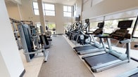 This single-level fitness center is designed to support a comprehensive workout experience, combining thoughtful architecture with a broad selection of professional-grade equipment. The main gym floor is defined by soaring double-height ceilings and an expansive wall of large-pane windows that flood the space with natural light while offering views of the surrounding outdoors. A large ceiling fan circulates air throughout, enhancing comfort during workouts. The cardio zone features a row of Matrix treadmills, elliptical trainers, and stair climbers, all equipped with individual screens and positioned to take advantage of the natural light. The strength training area includes a variety of Matrix selectorized machines, cable systems, free-weight benches, and a multi-functional rack, with floor-to-ceiling mirrors along one wall to maximize the sense of space. A dedicated free weights and functional training room provides dumbbell racks, stability balls, resistance bands, foam rollers, and a cable machine, with a wall-mounted television and direct access to the adjacent studio. The group fitness studio features hardwood flooring, full-wall mirrors, a mounted display screen branded with "Fitness on Demand," TRX suspension trainers, and natural light from roller-shaded windows, making it ideal for yoga, stretching, or guided classes. The overall aesthetic is clean and contemporary, with neutral tones, recessed lighting, and carpeted flooring throughout the main areas.