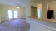 This single-floor space showcases an open-concept layout that seamlessly connects the kitchen and living areas. The kitchen features a raised granite-style breakfast bar with a clean white base, providing both a functional workspace and a natural divider between the cooking and living zones. A recessed nook adjacent to the bar offers additional storage or display potential.

The living area is finished with plush wall-to-wall carpeting in a neutral tone, lending warmth and comfort underfoot. Crown molding runs along the upper perimeter of the walls, adding a refined architectural detail throughout. A ceiling fan centered in the living space provides both lighting and air circulation.

Natural light filters into the room through a glass-paneled door along the far wall, brightening the space and offering a visual connection to the outdoors. The walls are painted in a soft, neutral tone that enhances the open and airy feel of the interior. A doorway visible in the background suggests additional rooms beyond the main living area. Overall, the space presents a clean, functional, and well-appointed interior suitable for comfortable everyday living.