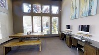 This business center offers a well-appointed, single-floor workspace that combines functional design with an open, airy atmosphere. The space is immediately distinguished by its soaring ceilings and an expansive bank of floor-to-ceiling windows, which flood the interior with generous natural light and provide views of the surrounding greenery and outdoor environment. The tall window arrangement, featuring both upper clerestory panes and larger lower panels with adjustable blinds, ensures consistent illumination throughout the day while maintaining privacy as needed. The main area is anchored by a sturdy rustic wood communal table paired with a matching bench, creating an inviting space for collaboration or informal meetings. Along one wall, a row of individual workstations equipped with desktop computers and dedicated desk space provides focused, private work areas suitable for task-oriented productivity. The neutral-toned carpet flooring and warm taupe walls contribute to a calm, professional ambiance. A piece of contemporary wall art adds a subtle creative accent, complementing the modern yet approachable aesthetic of the center. Overall, this business center balances open communal space with structured individual work areas, making it a versatile environment well-suited for a variety of professional activities.