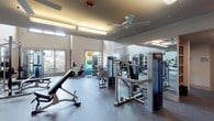 This two-story fitness center is thoughtfully designed to accommodate a wide range of workout disciplines across multiple dedicated zones. The layout spans two levels connected by a staircase, with a glass-railed mezzanine overlooking the main floor below, contributing to an open, airy atmosphere throughout the space.

The main level is divided into several functional areas. A cardio zone houses a row of Matrix treadmills and elliptical trainers positioned near large windows that invite natural light and views of the surrounding exterior. A separate strength training area features a comprehensive selection of Matrix cable machines, weight benches, barbells, and a full dumbbell rack, all set against floor-to-ceiling mirrors that enhance both visibility and the sense of space.

A dedicated cycling studio offers a row of indoor cycling bikes arranged before a wall of mirrors, with ceiling fans providing ventilation. An additional functional training area is equipped with medicine balls, resistance bands, stability balls, and a stationary recumbent bike, benefiting from soaring double-height ceilings and window views.

Throughout the facility, durable rubber flooring provides traction and impact absorption, while flush and linear ceiling lighting ensures consistent illumination. Ceiling fans are distributed across zones to maintain comfortable airflow during workouts.