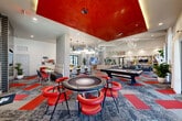 This contemporary amenity lounge is a thoughtfully designed, single-floor gathering space that seamlessly blends comfort, entertainment, and striking modern aesthetics. The open floor plan is anchored by a dramatic color palette of bold red, gray, and white, carried throughout the patchwork-style carpet and bold ceiling accents.

The lounge area features generous seating arrangements, including plush sofas, accent chairs, and communal tables, all bathed in natural light streaming through expansive floor-to-ceiling windows and glass doors. Soaring ceilings with architectural coffered details and a series of sculptural, contemporary chandeliers add visual drama throughout the space.

A full-length gourmet kitchen island with a gleaming white quartz countertop serves as a central social hub. The kitchen is equipped with stainless steel appliances, wall ovens, a full-size refrigerator, and a textured tile backsplash, all illuminated by a stunning abstract sculptural pendant light fixture.

The game and entertainment zone is defined by a bold lacquered red ceiling, housing a dedicated poker table with vibrant red seating, a billiards table, and a wall-mounted flat-screen TV. Smaller breakout nooks offer intimate seating for casual conversations. Lush indoor plants and curated artwork are displayed throughout, enhancing the vibrant, resort-inspired ambiance of this dynamic community space.
