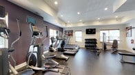 This single-floor fitness center is designed to provide a well-rounded exercise environment for users of all fitness levels. The space is outfitted with a lineup of cardio equipment, including elliptical trainers and treadmills, arranged to maximize floor space and allow for comfortable use. A dedicated free weights area features a multi-tiered dumbbell rack with a full range of weights, accompanied by a weight bench, making strength training convenient and accessible. A wall-mounted flat-screen television is positioned for easy viewing during workouts, adding an element of entertainment to training sessions. The interior is finished with a durable dark rubberized flooring suited for high-traffic exercise use, while the walls are painted in a clean two-tone scheme of white and deep purple, lending a motivating and modern aesthetic. Recessed lighting in the tray ceiling ensures the space is well-lit throughout. Multiple windows with blinds allow for natural light while maintaining privacy. Motivational fitness artwork adorns the walls, reinforcing the space's athletic atmosphere. Overall, the fitness center combines practical equipment layout with a clean, modern design to create an inviting and functional workout environment.