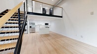 This contemporary two-story loft in Grand Rapids, Michigan offers a well-designed open-concept layout with modern finishes throughout. The main floor impresses with soaring ceilings, light hardwood flooring, and a dramatic floor-to-ceiling multi-pane window that floods the space with natural light and offers views of the surrounding streetscape. A striking black steel staircase with wood-toned treads and a cable railing system serves as both a functional and architectural focal point, leading up to the mezzanine level above.

The kitchen is sleek and modern, featuring white cabinetry, a central island with a clean white countertop, stainless steel appliances including a French door refrigerator and microwave, and in-unit stacked laundry. Recessed lighting illuminates the space efficiently throughout. The main floor also benefits from a ceiling fan, adding to the overall comfort of the open living area.

The upper mezzanine level, enclosed by a matching cable railing, provides additional flexible living space overlooking the main floor below. The full bathroom is finished in a clean, all-white palette with a vanity, tub-shower combination with a glass door, and matching light hardwood-style flooring, maintaining the cohesive modern aesthetic found throughout the entire space.
