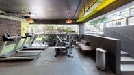 This single-level fitness center combines functional design with distinctive aesthetic touches to create an engaging workout environment. The space is anchored by polished concrete floors and dark charcoal walls and ceilings, lending it a modern, industrial character. A bold, colorful mural—reminiscent of Keith Haring's pop art style—serves as a striking focal point near the entrance, adding energy and personality to the interior.

The gym is equipped with a comprehensive selection of cardio machines, including treadmills, ellipticals, and stationary bikes, as well as a cable strength training station. Warm woven pendant lights are suspended from the ceiling, softening the industrial palette and providing an inviting ambiance. A vivid yellow-green accent panel on the ceiling adds a pop of contrast and visual interest.

One of the standout features of the space is its open-air connectivity to the outdoors. Large horizontal openings along one wall offer views of surrounding greenery, trees, and what appears to be a pool or outdoor lounge area, allowing natural light and fresh air to flow into the gym. Planters with tropical foliage line this open edge, reinforcing the connection to the outdoors.

A seating area with cushioned benches provides a place to rest, while a decorative laser-cut metal panel with a wave pattern adds a refined, artistic detail to the back wall. The overall atmosphere is modern, creative, and energizing.
