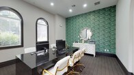This single-story space combines architectural elegance with refined interior design to create a polished and professional environment. The layout flows seamlessly between areas, connected by graceful arched doorways that frame views into a lounge area featuring a tray ceiling with recessed lighting and a statement chandelier. Arched windows with dark trim are a recurring architectural detail throughout, flooding the interior with natural light while adding a classic, upscale character.

The primary workspace is designed for both function and style, featuring sleek black granite desks paired with white and gold rolling chairs that lend a glamorous touch. A striking feature wall covered in an intricate emerald green malachite-patterned wallpaper serves as a bold focal point, complemented by a white ornate sideboard styled with decorative mirrors, candleholders, and gold accents. Recessed lighting throughout keeps the space well-lit, while neutral gray carpeted flooring with subtle patterning grounds the overall aesthetic. The lounge area beyond the archways offers comfortable seating and warm-toned walls, providing a welcoming transition within the space. The overall ambiance is sophisticated, visually dynamic, and thoughtfully curated.