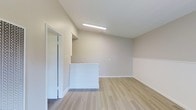 This single-floor unit offers a thoughtfully designed layout that maximizes its compact footprint. The main living area is characterized by light-toned hardwood-style flooring that runs throughout the space, lending warmth and visual continuity. Vaulted ceilings rise above the room, creating an enhanced sense of openness that extends the perceived volume beyond what the floor plan alone might suggest.

The walls are finished in a soft, neutral beige tone, contributing to a bright and calming ambiance. A flush-mounted linear light fixture provides consistent overhead illumination, ensuring the space remains well-lit. Clean white baseboards and trim offer a crisp contrast against the flooring and walls.

A white half-wall partition defines a functional zone within the space, which may serve as a small reception counter or divider, adding structure without enclosing the area. A mirrored door nearby reflects light and adds depth to the room. Light switch panels are positioned conveniently along the walls.

The overall aesthetic is clean, modern, and move-in ready, suited for a variety of uses. The minimalist design allows for flexible furnishing and personalization while maintaining an uncluttered, efficient atmosphere.