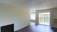 This single-floor space presents a clean, functional layout with a comfortable living area as its centerpiece. The room is finished with dark hardwood flooring that contrasts nicely against the neutral-toned walls, creating a warm and grounded aesthetic throughout. A textured popcorn ceiling adds a classic touch overhead, while a ceiling fan provides practical air circulation.

Natural light plays a significant role in the ambiance of this space. A large window fitted with horizontal blinds allows ample daylight to filter in, and a sliding glass door leads directly to a private balcony, offering outdoor views of surrounding greenery. The balcony access enhances the sense of openness and connection to the outdoors.

The living area also features a baseboard heating unit positioned beneath the window, ensuring efficient warmth during cooler months. A fireplace is partially visible along one wall, adding a cozy architectural detail to the room. The overall design is straightforward and neutral, making it an adaptable space well-suited for a variety of furnishing styles and personal preferences.