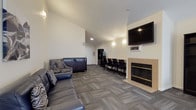 This single-floor community space is thoughtfully designed to serve as a versatile gathering and activity hub. The main lounge area is warm and inviting, anchored by a tile-surround fireplace with a wall-mounted flat-screen TV above it, making it an ideal spot for relaxation or casual entertainment. Comfortable seating is arranged throughout, including a navy blue leather sofa, patterned accent chairs, and side tables, all set atop multi-tone patterned carpet that adds texture and depth to the neutral, light-colored walls. Wall sconces provide soft ambient lighting, and abstract artwork accents the lounge area. Adjacent to the lounge, a dedicated workstation area features a row of task chairs and a long desk surface, offering a functional space for work or study. A dark bookshelf unit and a dining table with chairs provide additional utility and versatility. The fully equipped kitchen features warm honey-toned wood cabinetry, granite-style countertops, wood-look vinyl flooring, and black appliances including a range, microwave, refrigerator, and dishwasher. The kitchen transitions seamlessly into the carpeted common area, creating an open, connected flow throughout the space. Overall, the design balances comfort and functionality in a clean, modern aesthetic.