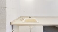 Located at 29-31 Upper Canada Dr in Toronto's St. Andrew-Windfields neighbourhood, this shared laundry facility offers a practical and efficiently organized space for residents. The room follows a long, narrow layout with light beige ceramic tile flooring throughout, contributing to a clean and easy-to-maintain environment. Bright fluorescent overhead lighting ensures the space is well-lit at all times, supplemented by a window at the far end that allows natural light to filter in.

A row of front-loading washing machines lines one wall, accompanied by a corresponding set of top-loading dryers, providing ample capacity for multiple users. On the opposite side, a generous continuous countertop offers dedicated space for folding laundry, with an under-counter utility sink featuring a chrome faucet and cabinet storage below — ideal for pre-treating garments or handwashing delicates. An information board near the entrance displays posted notices and guidelines for users.

The walls are finished in white, reinforcing the room's clean, functional aesthetic. A card-operated payment terminal is conveniently positioned near the entrance. The space also includes a small seating area with a chair beside the folding counter, allowing users to wait comfortably. Overall, this laundry facility is designed with practicality and user convenience at the forefront.