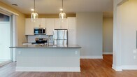 This well-designed single-floor residence showcases a thoughtful open-concept layout that seamlessly connects the kitchen and living areas. The kitchen serves as a functional centerpiece, featuring a large island with a dark countertop, an integrated sink, and elegant pendant lighting with glass globe fixtures. White shaker-style cabinetry lines the walls, complemented by a classic white subway tile backsplash and a full suite of stainless steel appliances, including a range, microwave, and refrigerator. Warm-toned hardwood-style flooring flows throughout the main living areas, creating a cohesive and inviting atmosphere. The spacious living area benefits from multiple windows and sliding glass door access, filling the space with natural light and views of surrounding greenery. Bedrooms are finished with plush neutral carpet, offering a comfortable retreat, with one bedroom featuring a wide window with outdoor views. Two bathrooms are present, each appointed with white subway tile, wood-look flooring, and navy blue vanities with brushed nickel hardware — one featuring a soaking tub and shower combination, and the other a walk-in shower.