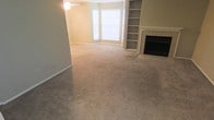 This single-story space centers around a generously sized living area finished with wall-to-wall carpet in a neutral beige tone, creating a warm and cohesive atmosphere throughout. A prominent fireplace with a tiled surround and classic white mantelpiece serves as a natural focal point along one wall, flanked by built-in shelving units that provide ample display and storage space. The shelving's integrated design adds a sense of architectural character to the room. A bay window arrangement along the adjacent wall allows natural light to filter in, brightening the interior and lending an open, airy quality to the space. Neutral warm-toned walls complement the flooring and contribute to the understated, move-in-ready aesthetic. A ceiling fan is centrally positioned overhead, offering both airflow and supplemental lighting. The layout flows openly, accommodating a variety of furniture arrangements. Overall, the space presents a functional and comfortable living environment with classic architectural details and practical built-in features.