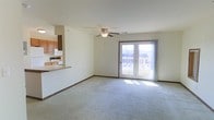 This single-floor residential space offers a comfortable and functional layout centered around an open-concept living and kitchen area. The spacious living room is finished with plush wall-to-wall carpeting and features a ceiling fan with light fixture, providing both comfort and practical overhead illumination. Neutral-toned walls and warm wood baseboards throughout lend the space a clean, inviting ambiance.

A highlight of the living area is the set of French doors that flood the room with natural light and open directly onto an outdoor deck, offering pleasant outdoor views and easy access to the exterior. The bright, airy feel created by these doors makes the living space feel open and connected to the outdoors.

The kitchen is positioned adjacent to the living area and is separated by a raised breakfast bar counter, creating a natural divide while maintaining an open flow between the two spaces. The kitchen is equipped with white appliances, including a refrigerator, microwave, and range, and features warm wood cabinetry that adds a traditional, homey character. The breakfast bar provides a convenient spot for casual dining or entertaining.

Overall, the space presents a practical and welcoming single-floor layout with thoughtful design elements that maximize natural light and functional living.