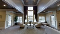 This single-floor space presents a refined and sophisticated lounge area defined by its dramatic architectural design. The most striking feature is the soaring ceiling height, accentuated by a coffered tray ceiling with dark painted insets that create a bold contrast against the crisp white crown molding and architectural trim. A pendant chandelier descends from the center, adding a touch of elegance.

Natural light floods the interior through an impressive floor-to-ceiling window arrangement at the far wall, flanked by tall, dark drape panels that frame the view and add a layer of visual depth. Additional side windows further enhance the brightness throughout the space. Columns with wainscoting detail line the walls, contributing to the classical aesthetic.

The seating area is centrally arranged, featuring cream-upholstered armchairs and a sofa grouped around a sleek glass-top coffee table, creating an inviting conversation area. Neutral-toned carpeting anchors the space, while a large potted plant introduces a natural element. The overall ambiance is polished, airy, and well-appointed, suited for formal gathering or reception use.