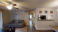 This single-story residence offers a practical and well-organized open-concept layout. The living area serves as the heart of the home, featuring dark hardwood-style flooring, a ceiling fan, and a textured ceiling. A large abstract canvas anchors one wall, adding a contemporary touch to the space. A hallway visible from the living area leads to additional rooms, with natural light filtering through at the far end, hinting at well-placed windows throughout the unit.

Adjacent to the living area, a kitchen pass-through counter creates a natural division between the two spaces while maintaining an open, connected feel. The kitchen is equipped with black appliances, including a refrigerator and microwave, and features light cabinetry. The pass-through design allows for easy interaction between the kitchen and living areas, making it well-suited for everyday living and casual entertaining.

The flooring transitions subtly between spaces, and neutral wall tones throughout provide a cohesive, understated backdrop. The layout is straightforward and functional, with each area flowing naturally into the next, creating a comfortable and livable environment.
