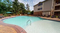 This outdoor amenity space is designed for relaxation and social gatherings, combining thoughtful architectural details with comfortable, resort-style features. At its centerpiece is a generously sized freeform swimming pool with clear, turquoise water, bordered by a classic brick coping and a paved pool deck. The deck accommodates multiple lounge chairs and a shaded umbrella area, making it ideal for sunbathing or unwinding outdoors. Mature trees and neatly maintained landscaping surround the perimeter, providing both privacy and a verdant backdrop.

Adjacent to the pool, a covered pavilion serves as the social hub of the space. Sturdy stone-clad columns support the overhead structure, lending a refined, upscale aesthetic. Inside the pavilion, a stone fireplace with a wall-mounted television creates a cozy focal point, complemented by plush wicker seating — including a sofa, armchair, and ottoman — all upholstered in neutral, patterned fabrics. The flooring throughout the pavilion and surrounding patio is composed of interlocking paver stones, adding texture and visual warmth.

A built-in grill station is conveniently positioned near the pool entrance, supporting outdoor dining and entertaining. Dining furniture is also available on the open patio, providing space for alfresco meals. Together, these elements create a cohesive, inviting outdoor retreat well-suited for both quiet relaxation and lively social events.