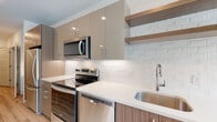 This well-designed single-floor unit offers a clean, modern aesthetic throughout, with light hardwood-style flooring and crisp white walls creating a bright, cohesive atmosphere. The open-concept kitchen is a standout feature, showcasing a two-tone cabinetry design that pairs warm wood-grain lower cabinets with sleek taupe upper cabinets. A textured white tile backsplash, quartz countertops, stainless steel appliances — including a refrigerator, range, microwave, and dishwasher — and an undermount sink with a contemporary faucet complete the well-appointed cooking space. Open floating shelves add a stylish, functional touch. The living area is generously proportioned, benefiting from natural light through large windows and a glass-paned entry door, with a ceiling fan adding comfort. A built-in desk nook offers a convenient workspace just off the kitchen. The bathroom features neutral tile flooring, a soaking tub and shower with subway tile surround, and a wood-cabinet vanity with a white countertop. The in-unit laundry area houses a stacked Whirlpool washer and dryer alongside wire shelving for additional storage. A dedicated hallway pantry with adjustable shelving rounds out the ample storage options found throughout this thoughtfully designed space.