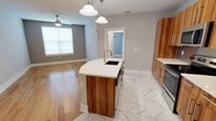 This well-designed single-floor space offers a seamless open-concept layout that connects the kitchen and living areas, creating a bright and functional environment. The kitchen is a standout feature, anchored by a curved central island topped with a crisp white countertop and equipped with an undermount sink and pendant lighting. Warm wood-toned cabinetry adds a natural, contemporary contrast to the neutral gray walls and light-colored surroundings. The kitchen flooring features large-format marble-look tile with subtle veining, lending an elegant touch underfoot, while the adjoining living area transitions to smooth hardwood flooring, adding warmth and continuity. Natural light filters in through a sizable window in the living area, enhancing the open and airy feel of the space. A ceiling fan is also present in the living area, offering both style and comfort. The kitchen is further equipped with stainless steel appliances, complementing the modern aesthetic. A doorway leads to an additional room, providing a clear sense of flow and connectivity throughout the layout. Overall, the space balances modern design with practical functionality in a clean, inviting atmosphere.