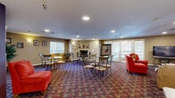 Located at 2390 Ridgeview Dr in Eau Claire, Wisconsin, this expansive single-floor community space offers a well-rounded mix of recreational and social amenities spread across a generously sized floor plan. The centerpiece of the facility is a large indoor swimming pool set beneath soaring vaulted ceilings clad in warm wood paneling. Arched windows and surrounding skylights flood the pool area with natural light, while a hot tub and poolside seating provide additional relaxation options. An entry vestibule with a natural wood ceiling and polished concrete floors serves as a welcoming transition into the facility. The open lounge and living area is warmly appointed with a striking stone fireplace, rich patterned carpeting, and a mix of dining tables, chairs, and comfortable seating. Bold accent walls in deep plum and warm gold tones add character throughout, complemented by recessed lighting and framed artwork. A dedicated television lounge offers leather seating and media viewing. The full kitchen features white cabinetry, a central island with a granite-style countertop, a full suite of appliances, and tile flooring — well-suited for catered events or community use. Together, these spaces create a versatile, welcoming environment designed for social engagement and active living.