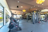 Located at 27665 Forbes Rd in Laguna Niguel, CA, this single-floor fitness center is a well-appointed facility designed to support a comprehensive workout experience. The space is bathed in abundant natural light, courtesy of expansive floor-to-ceiling windows that line the walls and offer views of the surrounding landscaped greenery — creating an open, energizing atmosphere throughout.

The cardio zone is generously equipped with multiple treadmills, stair climbers, and elliptical machines, all positioned along the windowed walls to maximize outdoor views during workouts. The strength training area features a full rack of dumbbells in varying weights, barbells, weight benches, and a multi-station cable machine, providing ample options for resistance training. A dedicated functional training zone includes a TRX suspension training rig, medicine balls, and other functional fitness tools.

Textured decorative wall panels and framed mirrors amplify the sense of space and add a refined aesthetic touch. A large flat-screen television is mounted prominently on the wall, offering entertainment during workouts. Overhead, distinctive sculptural pendant lights in a warm gold hue complement the recessed lighting to create a balanced, modern ambiance. Durable rubber flooring throughout ensures safety and comfort, making this fitness center both practical and visually appealing.