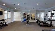 This dedicated fitness space is thoughtfully designed to accommodate a comprehensive workout experience within a single-floor layout. The room features neutral gray walls, crown molding, and a textured ceiling with recessed lighting that provides bright, even illumination throughout. Carpeted flooring adds comfort underfoot, making the space suitable for stretching and floor exercises. The workout area is outfitted with a variety of fitness equipment, including cardio machines, a stationary exercise bench, and a stacked set of exercise balls in varying sizes. A balance dome trainer is also visible, catering to stability and core workouts. Large wall-mounted mirrors are strategically placed to expand the sense of space and allow users to monitor their form during exercise. Natural light filters in through multiple windows with white trim, enhancing the open and airy ambiance of the room. A glass partition element adds a modern architectural detail, subtly defining areas within the space. The overall design strikes a balance between functionality and comfort, creating an inviting environment well-suited for both light and moderate fitness routines.