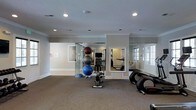 This dedicated fitness space is thoughtfully designed to accommodate a comprehensive workout experience within a single-floor layout. The room features neutral gray walls, crown molding, and a textured ceiling with recessed lighting that provides bright, even illumination throughout. Carpeted flooring adds comfort underfoot, making the space suitable for stretching and floor exercises. The workout area is outfitted with a variety of fitness equipment, including cardio machines, a stationary exercise bench, and a stacked set of exercise balls in varying sizes. A balance dome trainer is also visible, catering to stability and core workouts. Large wall-mounted mirrors are strategically placed to expand the sense of space and allow users to monitor their form during exercise. Natural light filters in through multiple windows with white trim, enhancing the open and airy ambiance of the room. A glass partition element adds a modern architectural detail, subtly defining areas within the space. The overall design strikes a balance between functionality and comfort, creating an inviting environment well-suited for both light and moderate fitness routines.