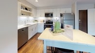 This single-floor residence combines modern finishes with a thoughtfully designed layout across several well-defined living spaces. The kitchen is a standout feature, showcasing white shaker cabinetry extending to the ceiling, a mosaic tile backsplash, stainless steel appliances, and a quartz-topped island with bar seating — all set atop rich hardwood flooring that flows throughout the main living areas.

The living room is bright and welcoming, with large sliding glass doors that open directly to a private patio, framing views of lush landscaping. Track lighting and neutral walls create a versatile backdrop for the space.

The residence offers multiple bedrooms, each carpeted for comfort. The primary bedroom connects to an en-suite bathroom featuring a white vanity with quartz countertops, an illuminated mirror, and large-format tile flooring. A second full bathroom echoes the same clean aesthetic with ample storage cabinetry. A dedicated in-unit laundry closet with a full-size washer and dryer adds everyday convenience.

Throughout, the design balances contemporary style with functional living, presenting a cohesive and polished interior.