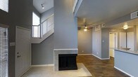 This two-story space in Arlington, Texas offers an open and airy layout defined by soaring ceilings that amplify the sense of vertical space. The main living area centers around a striking floor-to-ceiling fireplace framed in granite-style tile, serving as a bold architectural focal point. Gray-toned walls carry throughout, lending a cohesive and modern aesthetic to the interior.

The living area features a combination of carpet and hardwood-style flooring, adding warmth and texture underfoot. A ceiling fan with integrated lighting provides both comfort and ambient illumination, while natural light filters in through windows positioned at both floor levels, brightening the space throughout the day.

The staircase, visible from the main living area, features a windowed landing that draws in additional natural light and adds an element of openness to the vertical transition between floors. HVAC vents are integrated into the upper wall, ensuring efficient climate control throughout the space. The overall ambiance is clean, neutral, and functional, making it well-suited for a variety of furnishing and design styles.