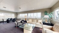 This well-appointed single-floor space is thoughtfully designed to accommodate both relaxed gatherings and formal meetings. The main lounge area is generously proportioned, featuring plush cream-colored sofas and armchairs, including a supple leather accent chair, all arranged around a glass-top coffee table. A striking natural stone fireplace serves as a central focal point, complemented by a wall-mounted television set against blue-painted wood paneling, adding a warm and inviting character to the room. Polished floor lamps provide ambient lighting throughout, while lush potted plants bring a touch of greenery to the interior. The space is carpeted throughout in a dark patterned carpet that lends a classic, refined feel underfoot. Large sliding windows wrap around multiple walls, flooding the interior with natural light and offering expansive panoramic views of the surrounding skyline and open sky, with access to an outdoor terrace visible from the lounge. A dedicated meeting or dining area features a glass-top conference table surrounded by sleek dark leather chairs, ideal for formal discussions or seated gatherings. The neutral taupe wall tones and drop-panel ceiling tie the entire space together, creating a cohesive and professional yet comfortable ambiance throughout.