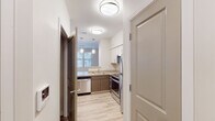 This single-floor residence features a contemporary, neutral design throughout. The kitchen is well-appointed with stainless steel appliances, including a refrigerator, range, microwave, and dishwasher. Dual-tone cabinetry pairs warm wood-finish lower cabinets with crisp white uppers, complemented by light countertops. Pendant lighting and a window bring natural light into the space, while light wood-style flooring extends through the kitchen and hallway, adding warmth and visual continuity.

The bedroom is generously sized, featuring plush carpeting and multiple windows that allow ample natural light and outdoor views. A dedicated walk-in closet with built-in shelving provides practical storage. The bathroom is finished in a clean, functional style with a single-sink vanity topped with a white countertop, warm wood cabinetry, a large mirror, and a walk-in shower. Tile flooring and a bright vanity light fixture complete the space.

The 3D floor plan reveals a compact, efficiently arranged layout that connects the main living areas, kitchen, bedroom, bathroom, and an in-unit laundry area through a central hallway, maximizing the use of available space.