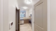 This single-floor residence features a contemporary, neutral design throughout. The kitchen is well-appointed with stainless steel appliances, including a refrigerator, range, microwave, and dishwasher. Dual-tone cabinetry pairs warm wood-finish lower cabinets with crisp white uppers, complemented by light countertops. Pendant lighting and a window bring natural light into the space, while light wood-style flooring extends through the kitchen and hallway, adding warmth and visual continuity.

The bedroom is generously sized, featuring plush carpeting and multiple windows that allow ample natural light and outdoor views. A dedicated walk-in closet with built-in shelving provides practical storage. The bathroom is finished in a clean, functional style with a single-sink vanity topped with a white countertop, warm wood cabinetry, a large mirror, and a walk-in shower. Tile flooring and a bright vanity light fixture complete the space.

The 3D floor plan reveals a compact, efficiently arranged layout that connects the main living areas, kitchen, bedroom, bathroom, and an in-unit laundry area through a central hallway, maximizing the use of available space.