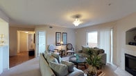 This single-floor residence centers around a thoughtfully designed living room that serves as the heart of the space. The living room is generously sized, offering ample room for multiple seating arrangements, including plush upholstered sofas and accent chairs arranged around a circular glass coffee table. Neutral beige tones on the walls and carpet create a warm, inviting ambiance throughout.

A textured ceiling with a classic semi-flush light fixture provides soft overhead illumination, complemented by a traditional floor lamp positioned near a pair of framed botanical wall art pieces. The accent chairs, featuring a mix of styles, add visual interest and a touch of elegance to the seating area.

Natural light filters into the room through a window dressed with vertical blinds, brightening the space and offering a view of the outdoors. Indoor potted plants, including a striking tropical specimen in the foreground, introduce organic color and life to the interior. A doorway visible to the left suggests additional rooms beyond, with hardwood-style flooring transitioning at the entryway. The overall atmosphere is calm, polished, and comfortable.