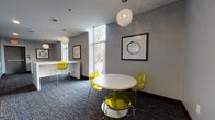 Located at 1449 S Church St in Charlotte's South End neighborhood, this single-floor commercial space offers a polished, modern environment designed for professional use. The layout includes a well-appointed conference room featuring a rectangular white table surrounded by white ergonomic chairs, a wall-mounted flat-screen display, recessed lighting, and framed artwork — ideal for meetings and presentations.

A lounge and collaborative area anchors the main gathering space, furnished with round white tables and distinctive yellow translucent chairs. Sculptural globe pendant lights hang overhead, complementing the textured gray walls and dark patterned carpeting throughout. Framed abstract artwork adds visual interest and a refined aesthetic.

Large floor-to-ceiling windows and tall vertical panes flood the space with natural light and provide street-level views of the surrounding tree-lined neighborhood. A built-in white counter desk near the windows offers an additional workstation, fitted with matching yellow accent chairs.

A central corridor connects the various zones, lined with sleek glass-panel partitions that enhance the open, airy feel while defining individual spaces. The overall design strikes a balance between collaborative openness and functional privacy, making it a versatile professional environment suited for office, creative, or business use.