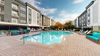 This outdoor amenity space at 3450 Miller Dr in Chamblee, Georgia, centers around a generously sized, freeform resort-style swimming pool with crystal-clear blue water. The pool area is framed by a broad brick-paved deck that provides ample room for relaxation and recreation. Surrounding the main pool is a dedicated wading area, offering a shallower option suitable for a range of users.

The deck is well-furnished with a mix of cushioned lounge chairs, chaise lounges, and upholstered seating in a signature teal accent color, arranged to maximize both sunbathing and social interaction. Mature trees and neatly maintained landscaping line the perimeter, providing natural greenery and partial shade throughout the space.

An adjacent outdoor patio area extends the amenity footprint and features café-style table seating with a market umbrella, alongside comfortable lounge furniture, creating a casual gathering spot. This patio connects to a covered structure with an upper-level terrace, adding vertical dimension to the outdoor environment.

The entire courtyard is enclosed by a black iron fence, ensuring privacy and security. The multi-story residential buildings surrounding the space provide an urban backdrop, while open sky above allows abundant natural light throughout the day.