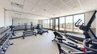 This fitness center is a compact yet thoughtfully outfitted exercise space designed to support a full range of workout routines. The room features a drop ceiling with recessed panel lighting and a neutral two-tone wall treatment — light gray on the upper half and a deeper sage green on the lower — creating a clean, focused environment for training.

The space is well-stocked with a variety of equipment, including Peloton-style stationary bikes with integrated screens, an elliptical trainer, a treadmill, and a rowing machine. A dedicated strength training area features a tiered dumbbell rack with a comprehensive set of weights, kettlebells, and barbells, along with weight benches to support resistance exercises. Yoga mats and resistance bands are neatly stored on wall-mounted racks near the entrance.

A standout feature of this fitness center is the wall of large floor-to-ceiling windows and sliding glass doors that flood the interior with abundant natural light and open directly onto an outdoor terrace. From this vantage point, sweeping city skyline views are visible, making workouts an immersive and energizing experience. The terrace itself includes outdoor seating, providing a space to cool down or relax post-workout. The overall ambiance is bright, modern, and functional — ideal for a focused fitness routine.