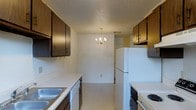 This two-floor space includes a galley-style kitchen that serves as a central feature of the home. The kitchen is lined on both sides with dark brown upper cabinetry, providing ample storage, while white lower cabinetry and light-toned countertops create a clean contrast. A double-basin stainless steel sink is positioned along one wall, accompanied by a white dishwasher and a white electric range on the opposite side. A full-size white refrigerator anchors the end of the kitchen run. The textured ceiling adds a subtle visual element throughout the space. The kitchen flows directly into a dining area, which is illuminated by a brass chandelier with candle-style lighting, giving the space a traditional touch. The light-colored flooring carries consistently from the kitchen into the dining area, reinforcing a sense of continuity. A closet is visible at the rear of the dining space, adding practical storage. The overall layout is efficient and straightforward, designed to maximize functionality while maintaining a cohesive, classic aesthetic across both levels of the home.