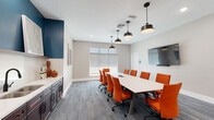 Located at 8710 SW 71st Ave in Ocala, Florida, this single-floor meeting space is thoughtfully designed to support professional gatherings and collaborative work sessions. The centerpiece of the space is a generously sized conference room featuring a sleek white rectangular table surrounded by vibrant orange ergonomic chairs, providing comfortable seating for a full group. Pendant lighting in a matte black finish hangs overhead, complementing the neutral gray wall-to-wall carpeting and light gray walls that give the room a clean, contemporary feel. A bold teal accent wall anchors one end of the room, adding visual depth and a pop of color that ties the design together. A wall-mounted flat-screen display serves as a dedicated presentation surface, while large windows along one wall allow natural light to filter in, offering views of the surrounding landscape. Adjacent to the main conference area is a well-appointed kitchenette featuring rich dark wood cabinetry, white quartz countertops, a double stainless steel sink with a matte black faucet, and upper shelving set against the same teal accent wall. A smaller breakout area with its own table and orange seating provides a more intimate setting for one-on-one meetings or focused work. Decorative artwork adds a finishing touch throughout, creating a polished and inviting professional environment.