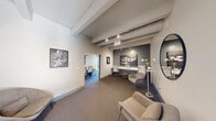 This thoughtfully designed single-floor commercial space combines contemporary aesthetics with industrial architectural elements to create a polished and functional environment. Exposed structural beams run across the elevated ceilings, complemented by track lighting that illuminates the space with a warm, focused glow. A striking sputnik-style chandelier serves as a standout centerpiece in the reception area, adding a sculptural quality to the overall design.

The layout flows naturally from a welcoming lounge area, furnished with modern upholstered seating, curated side tables, and a large round mirror that enhances the sense of openness. Carefully selected artwork adorns the walls throughout, reinforcing a gallery-like atmosphere. A dedicated workstation area features built-in desk surfaces set against a dark accent wall, creating a focused and productive zone.

Interior windows with teal-accented frames connect the spaces visually while maintaining distinct zones for collaboration and privacy. A separate conference room equipped with ergonomic seating is visible through open doorways. Floor-to-ceiling exterior windows in select areas invite natural light and offer views of the surrounding streetscape. Neutral carpet flooring and a soft, cohesive color palette of grays, whites, and teal accents tie the entire space together seamlessly.