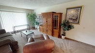 This single-floor residence offers a comfortable and functional layout across several distinct areas. The living room serves as the central gathering space, featuring plush carpeting, a textured ceiling, and a large window with vertical blinds that allows for generous natural light. A substantial wooden cabinet unit anchors the room, complemented by leather seating and a glass-top coffee table, creating a warm and inviting atmosphere.

Adjacent to the living room is a compact dining area, finished with the same neutral tones and textured ceiling. A round glass-top table with ornate wrought-iron chairs provides an intimate setting for meals.

The galley-style kitchen is efficiently designed with warm wood cabinetry lining both walls, offering ample storage. Light-toned countertops, a tile backsplash, and light flooring complete the space, which also benefits from a window providing natural ventilation.

The bedroom is a restful retreat with carpeted floors, a window for natural light, and a closet for storage. A connected hallway leads conveniently to the bathroom, which features clean white tile wainscoting, a vanity with a large mirror, and a well-lit Hollywood-style light bar above.

A secondary room functions as a flexible den or office space, with a window, hardwood-style flooring, and enough room to accommodate a desk, seating, and additional furnishings.
