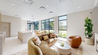 Located at 21580 Old Dixie Hwy in Miami, FL, this single-floor amenity lounge offers a thoughtfully designed open-plan layout that seamlessly blends comfort and modern sophistication. The space is anchored by expansive floor-to-ceiling windows along the exterior wall, flooding the interior with abundant natural light and framing lush greenery and outdoor views beyond.

The lounge area features plush curved seating, including a large sofa and rounded accent chairs in warm earth tones, arranged around a sculptural white marble coffee table. A dedicated entertainment zone includes a wall-mounted flat-screen TV set above a sleek linear fireplace, flanked by warm wood slat paneling that adds texture and visual warmth. A tall fiddle-leaf fig plant completes this cozy corner.

The open kitchenette is finished with light-toned cabinetry, a generous island with a crisp white countertop, a sink, and built-in open shelving displaying curated decorative accents. Adjacent to the kitchen, a casual dining area accommodates a natural wood table with woven chairs, bathed in sunlight streaming through the large windows.

Large-format light tile flooring, a recessed-lit drop ceiling, and a neutral warm palette throughout create a cohesive, airy ambiance that feels both inviting and refined — ideal as a communal gathering space.