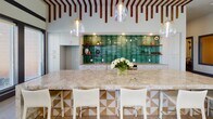 This thoughtfully designed single-floor amenity space combines modern aesthetics with functional gathering areas. The expansive main lounge impresses with a coffered ceiling accented by warm ambient lighting, a striking large-scale textured wall feature, and light wood-look tile flooring that runs throughout. Comfortable seating arrangements anchor the space, while floor-to-ceiling windows along the perimeter flood the interior with natural light and provide views of the surrounding outdoor areas.

The game room is a standout feature, outfitted with a shuffleboard table and a full-size pool table beneath an eye-catching geometric light fixture. An industrial-style glass garage door opens directly to an outdoor patio, seamlessly blending indoor and outdoor living.

The kitchen area showcases soaring ceilings with bold exposed wood beam detailing, a generous marble-topped island with seating, and a vibrant emerald-green tile backsplash that serves as a dramatic focal point. Pendant globe lights hang above the island, while white cabinetry, open wood shelving, and large windows complete the bright, contemporary look.

Throughout the space, wide glass-panel doors connect distinct zones, creating an open and flowing layout that is ideal for community gatherings and social events.