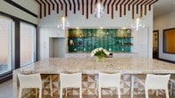 This thoughtfully designed single-floor amenity space combines modern aesthetics with functional gathering areas. The expansive main lounge impresses with a coffered ceiling accented by warm ambient lighting, a striking large-scale textured wall feature, and light wood-look tile flooring that runs throughout. Comfortable seating arrangements anchor the space, while floor-to-ceiling windows along the perimeter flood the interior with natural light and provide views of the surrounding outdoor areas.

The game room is a standout feature, outfitted with a shuffleboard table and a full-size pool table beneath an eye-catching geometric light fixture. An industrial-style glass garage door opens directly to an outdoor patio, seamlessly blending indoor and outdoor living.

The kitchen area showcases soaring ceilings with bold exposed wood beam detailing, a generous marble-topped island with seating, and a vibrant emerald-green tile backsplash that serves as a dramatic focal point. Pendant globe lights hang above the island, while white cabinetry, open wood shelving, and large windows complete the bright, contemporary look.

Throughout the space, wide glass-panel doors connect distinct zones, creating an open and flowing layout that is ideal for community gatherings and social events.