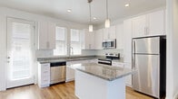 This two-story, 2-bedroom, 3-bathroom home is thoughtfully designed with a clean, modern aesthetic and consistent finishes throughout. The main floor combines the kitchen and living area into an open, light-filled space. The kitchen is a standout, featuring white shaker-style cabinetry, granite countertops, a central island with pendant lighting, stainless steel appliances including a dishwasher, range, microwave, and refrigerator, and warm hardwood flooring. Large windows with white blinds allow generous natural light into both the kitchen and the adjoining living area. A staircase with carpeted treads connects both floors, flanked by a window that brightens the transition. The upper level houses both bedrooms, each carpeted and featuring ceiling fans and windows with outdoor views. The primary bedroom is generously sized with direct access to a bathroom. The three bathrooms are finished with granite countertops, white cabinetry, framed mirrors, and brushed nickel or gold fixtures. One bathroom includes a full tub-shower combination with a window above. A hallway and closet on the upper level add practical storage and circulation. Throughout the home, recessed lighting, neutral white walls, and light hardwood floors on the main level create a bright, cohesive ambiance.