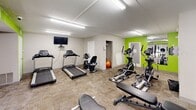 Located at 2 Colony Boulevard in Wilmington, Delaware, this fitness center offers a well-equipped exercise space designed to support a variety of workout routines. The room is finished with neutral-toned carpet flooring and bright overhead fluorescent lighting, creating a clean and functional environment. A bold lime-green accent wall adds a vibrant, energizing touch to the otherwise neutral color palette of white and gray walls.

The exercise room is outfitted with a comprehensive selection of cardio equipment, including multiple treadmills and elliptical machines, as well as a stationary bike for lower-impact training. A multi-function cable resistance machine and a weight bench provide strength training options. An exercise ball rounds out the available equipment. A large wall-mounted mirror spans one side of the room, enhancing the sense of space and allowing users to monitor their form during workouts. A wall-mounted flat-screen television is positioned for easy viewing from cardio equipment.

Natural light filters in through windows that offer views of the surrounding outdoor greenery, while a glass-panel door provides direct exterior access. The space also features a security camera, a safety information board, and a defibrillator unit mounted on the wall, underscoring a commitment to user safety. This fitness center delivers a practical and motivating environment for everyday exercise.