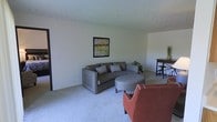 This single-floor space features an open living area designed for comfortable, everyday use. The living room is carpeted in a light neutral tone, creating a clean and inviting foundation throughout the space. A curved gray sofa serves as the centerpiece of the seating area, complemented by patterned throw pillows, a round upholstered ottoman, and a rust-colored accent chair that adds a warm pop of color. A framed abstract artwork adorns the white walls, adding a subtle decorative touch. The textured popcorn ceiling adds a classic character to the room. Natural light filters into the space, contributing to its bright and airy atmosphere. A tall bar-height table with stools is positioned toward the back of the room, offering a casual dining or workspace option. A small potted plant near the seating area introduces a touch of greenery. A doorway visible on the left side of the living area leads to additional rooms, suggesting a practical and straightforward floor plan. Overall, the space presents a functional, well-arranged layout with a relaxed and homey ambiance.