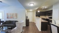 This single-floor residence offers a thoughtfully designed open-concept layout that connects the living area and kitchen in one cohesive, flowing space. Rich, dark hardwood-style flooring runs throughout, lending warmth and visual continuity to the interior.

The living area is welcoming and comfortably scaled, providing ample room for seating and relaxation. A front entryway with a clean white door anchors the space, offering a natural focal point upon entry and contributing to the bright, airy feel of the interior.

The kitchen is a standout feature, showcasing dark espresso cabinetry that creates a bold contrast against the light-toned walls and granite-style countertops. A peninsula with bar seating separates the kitchen from the main living area, making it ideal for casual dining or entertaining. Pendant lighting above the peninsula adds a modern, polished touch, while track lighting overhead ensures the kitchen is well-illuminated. Full-size appliances, including a refrigerator and range, are integrated seamlessly into the cabinetry.

Flush-mount ceiling fixtures provide consistent, even lighting throughout the space. The neutral wall palette enhances the sense of openness and serves as a versatile backdrop. Overall, the space presents a clean, modern aesthetic that balances functionality with style.