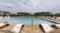 This single-story amenity space in Panama City, Florida, is designed to serve as a central outdoor retreat for its surrounding residential community. The centerpiece is a generously sized, curved swimming pool with crystal-clear blue water, accessible via stainless steel entry rails and complemented by a broad stone-tiled sundeck. The deck is furnished with rows of lounge chairs and distinctive white sculptural sun loungers positioned at the pool's edge, offering ample space for relaxation under the Florida sky.

Adjacent to the pool is a covered pavilion that functions as an open-air lounge area. The pavilion features panel-accented walls in neutral tones, recessed lighting, and a ceiling fan to promote airflow during warm months. White outdoor seating arrangements with teal accent cushions are organized around a central coffee table, creating a comfortable gathering space. Expansive doorways and windows connect the interior to the pool deck, allowing natural light to flow freely throughout.

A columned covered walkway extends along the building's exterior, providing shaded passage between the indoor amenity spaces and the pool area. Landscaped planters and fencing frame the perimeter, adding greenery and definition to the space. The overall aesthetic is clean, coastal, and modern, well-suited to the warm climate of the Florida Panhandle.
